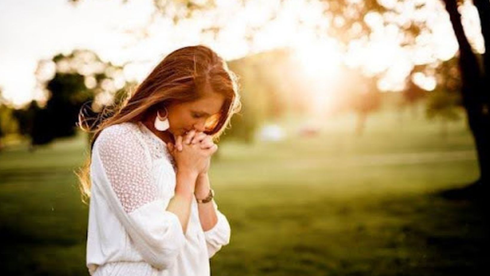 A young woman in a sunlit park with hands clasped in prayer.